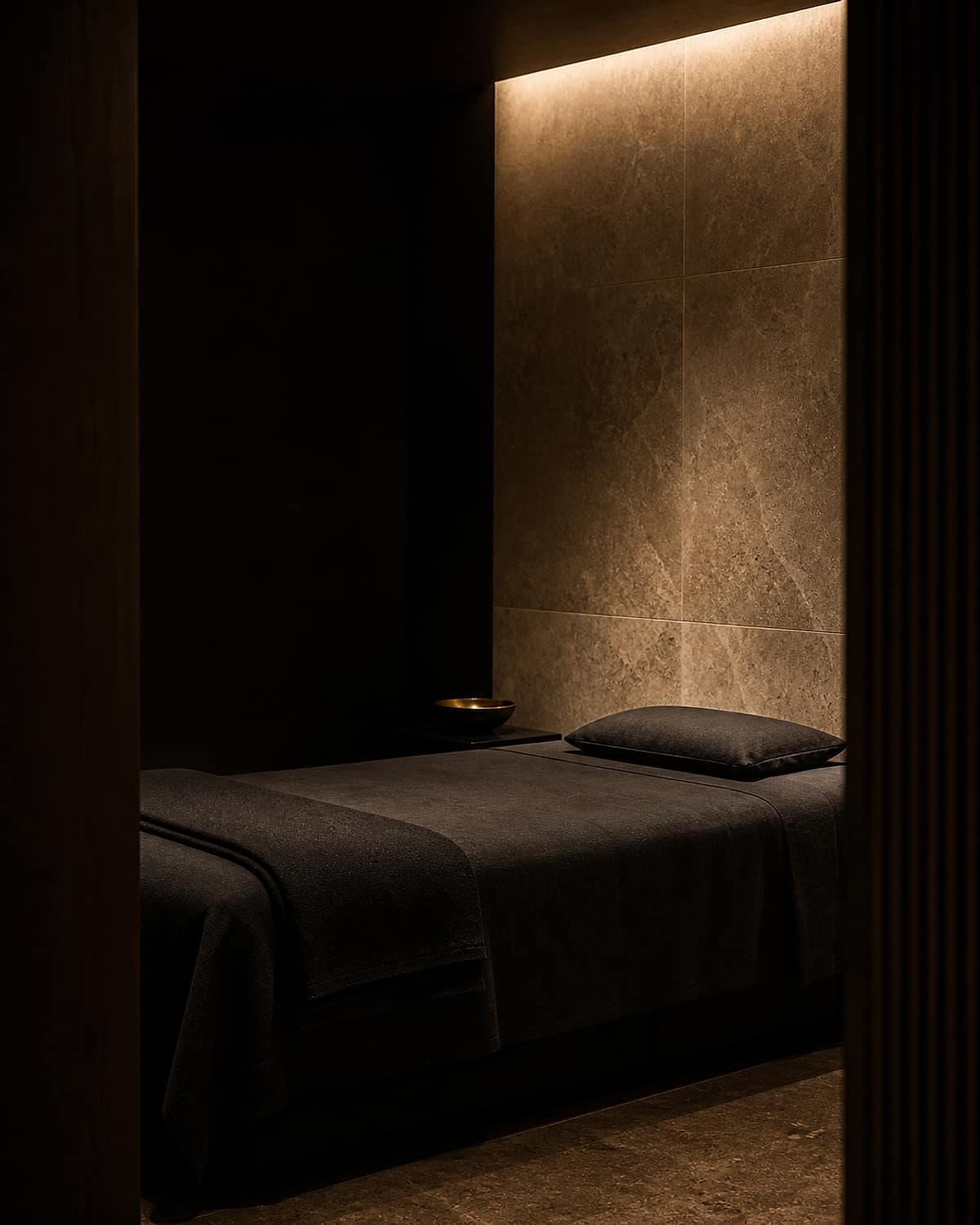 A minimal treatment room in low warm light, stone wall and dark linen